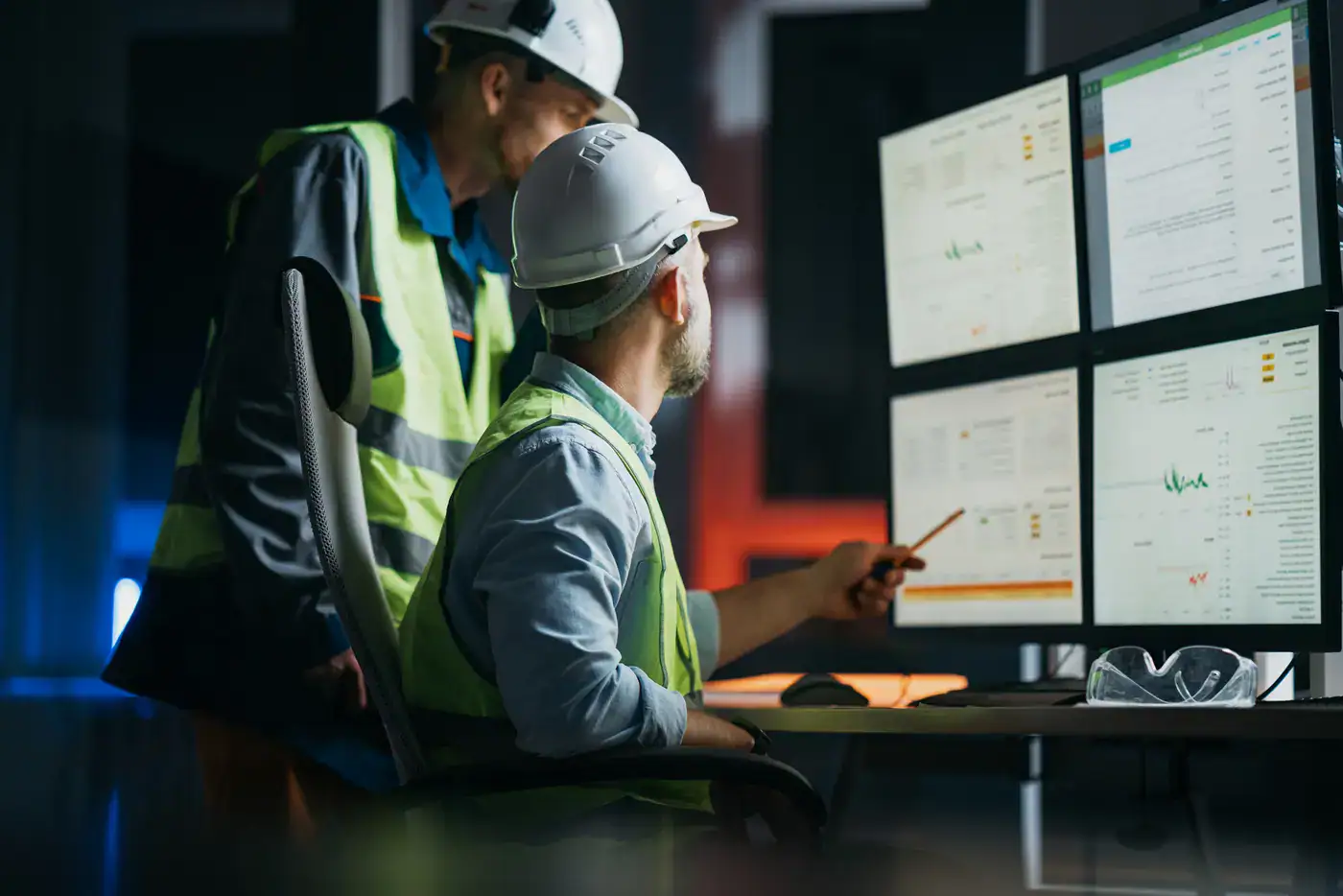 Industrial workers wearing hard hats and safety vests review operational data on multiple computer monitors in a control room.