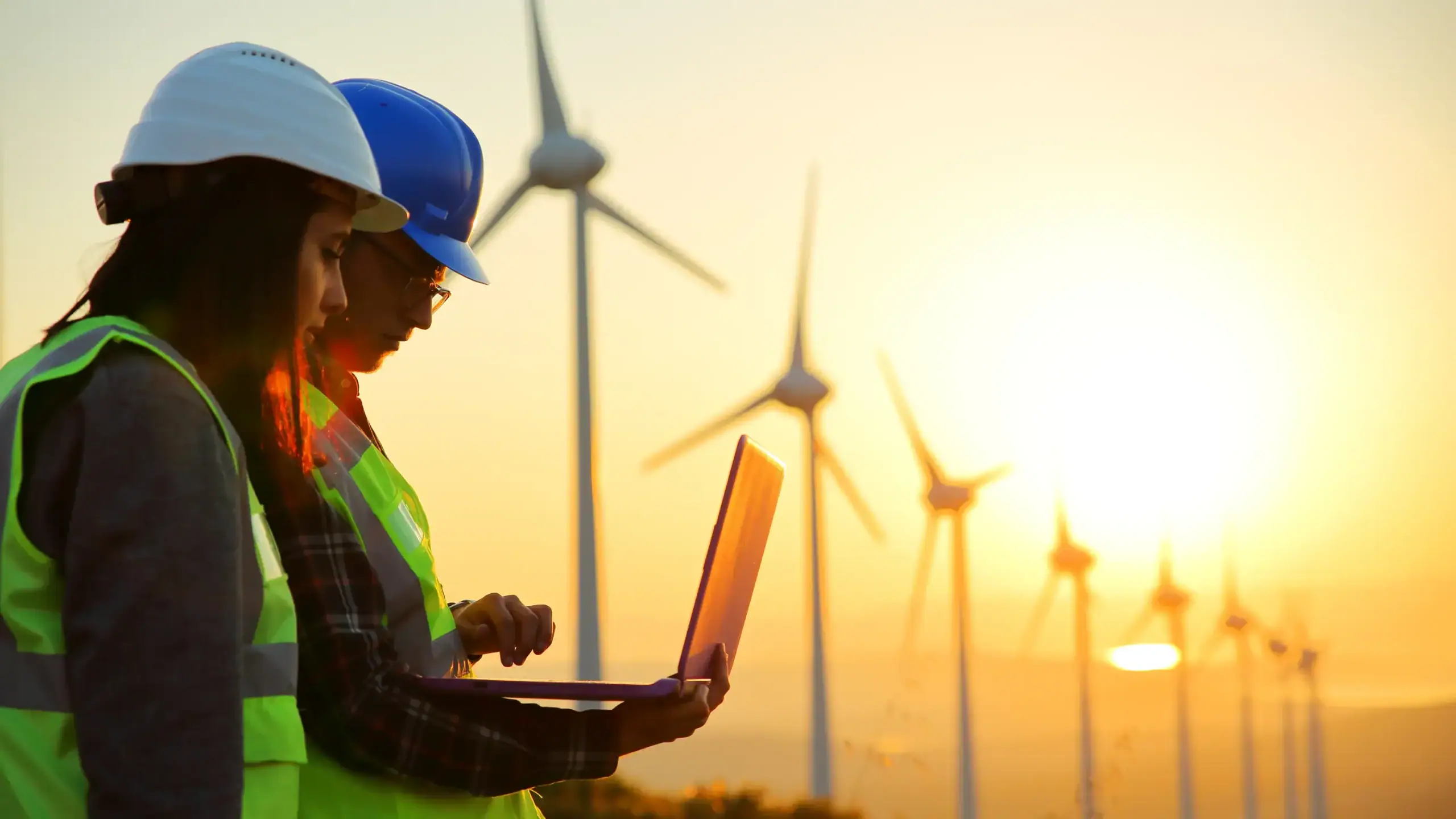 Technician using a laptop to monitor wind turbines at a renewable energy facility during sunset.