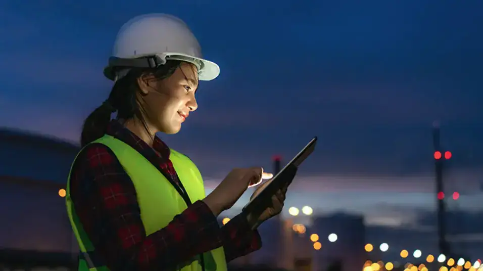 Worker in a hard hat and safety vest using a tablet to monitor operations at an industrial site during evening hours.