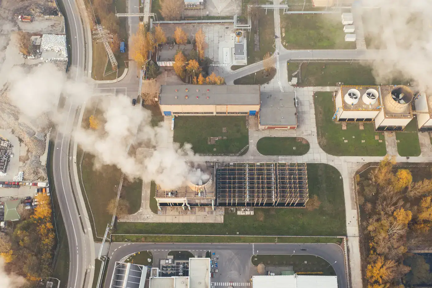 Aerial view of an industrial facility with buildings, roads, and visible steam or emissions rising from multiple structures.