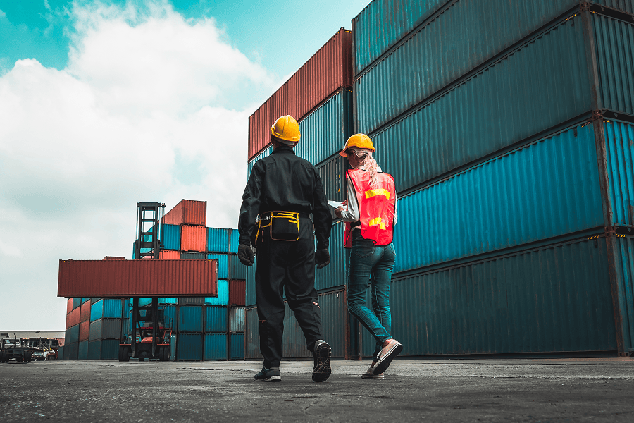 A maritime cargo container depot stacked with shipping containers, with two logistics workers walking by the yard