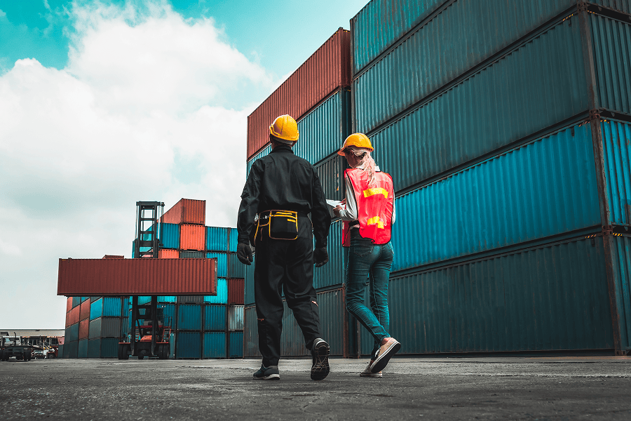 A maritime cargo container depot stacked with shipping containers, with two logistics workers walking by the yard
