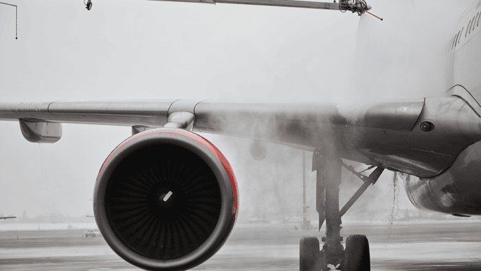 Commercial aircraft undergoing de-icing on an airport tarmac, with spray equipment applying fluid to the wing and engine area.