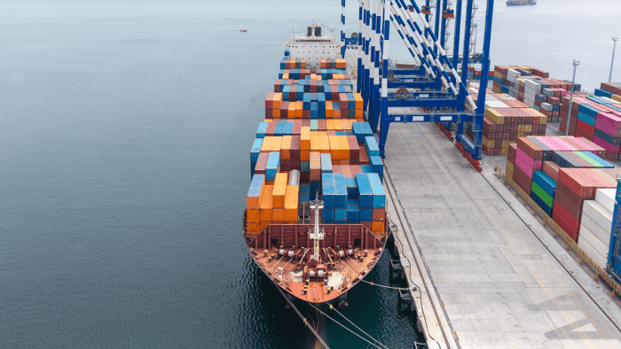 Container ship docked at a cargo terminal with stacked shipping containers and port cranes.