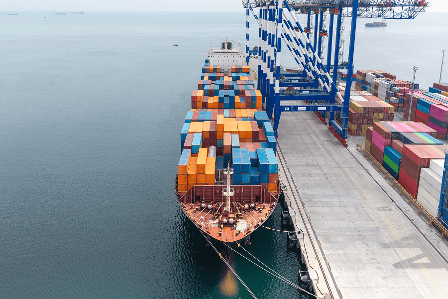 Container ship docked at a cargo terminal with stacked shipping containers and port cranes.