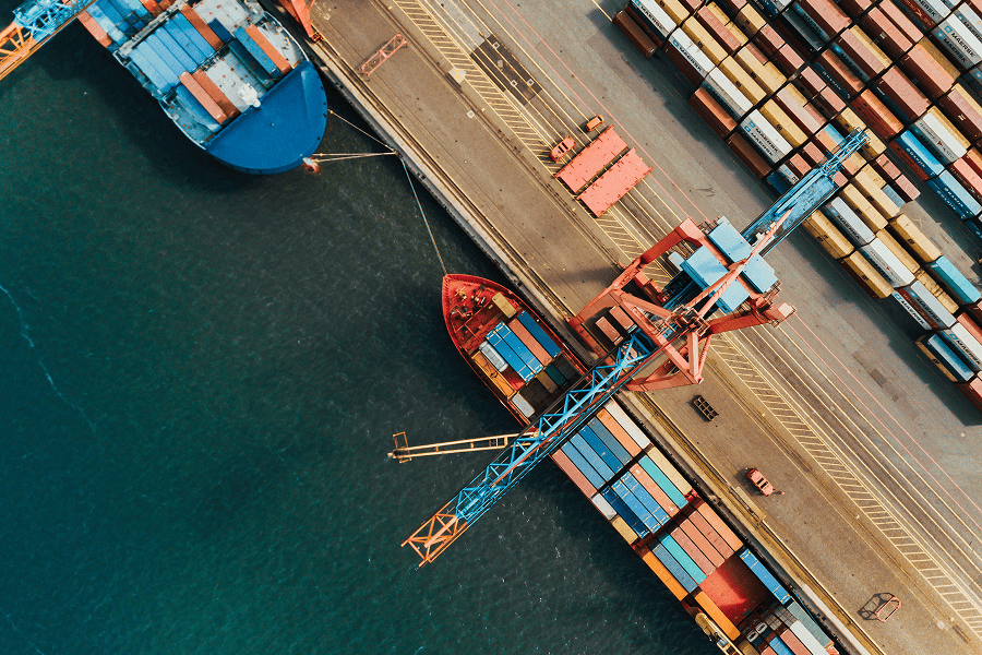 A commercial cargo vessel docked at a busy port terminal with an industrial container crane loading freight