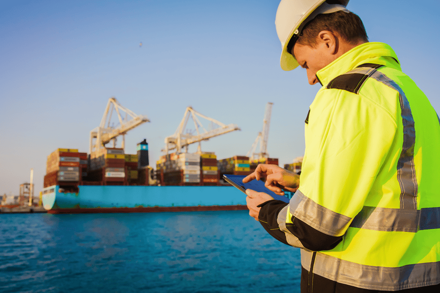 A marine engineer reviewing data on a digital tablet with a large cargo vessel docked in the background.