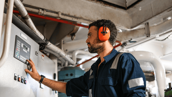 Industrial technician operating a control panel while wearing hearing protection inside a mechanical equipment room.