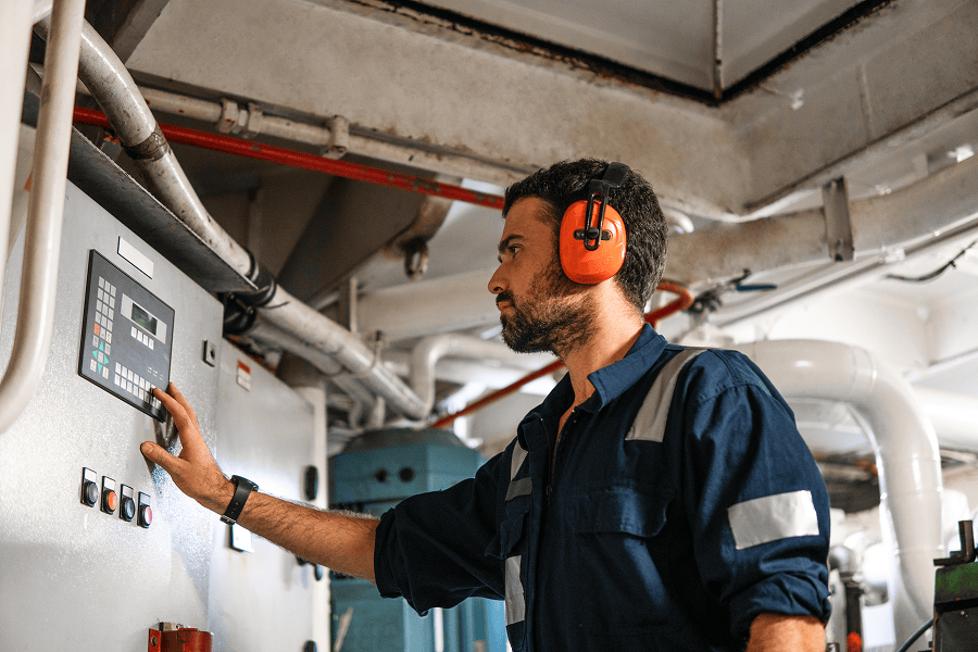 Industrial technician operating a control panel while wearing hearing protection inside a mechanical equipment room.