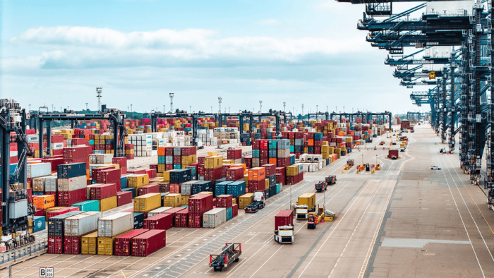 Busy container terminal with stacks of shipping containers, cargo handling equipment, and transport vehicles.