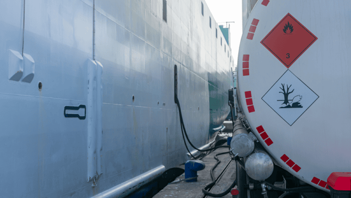 A detailed close-up view of a commercial tanker truck connection hose refueling a maritime vessel at a port.