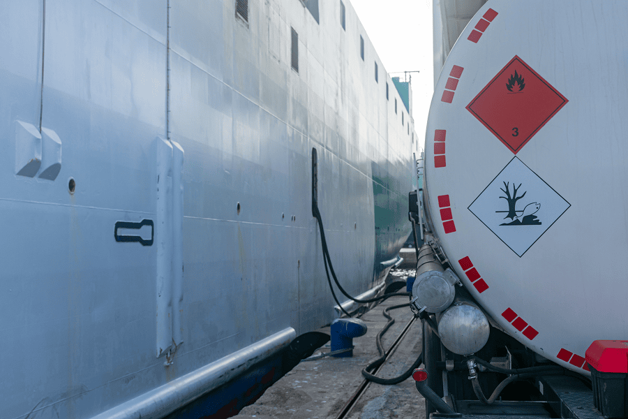 A detailed close-up view of a commercial tanker truck connection hose refueling a maritime vessel at a port.