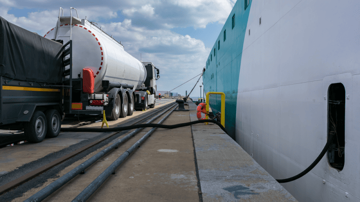 A commercial tanker truck parked at a port terminal pier actively refueling a docked maritime vessel