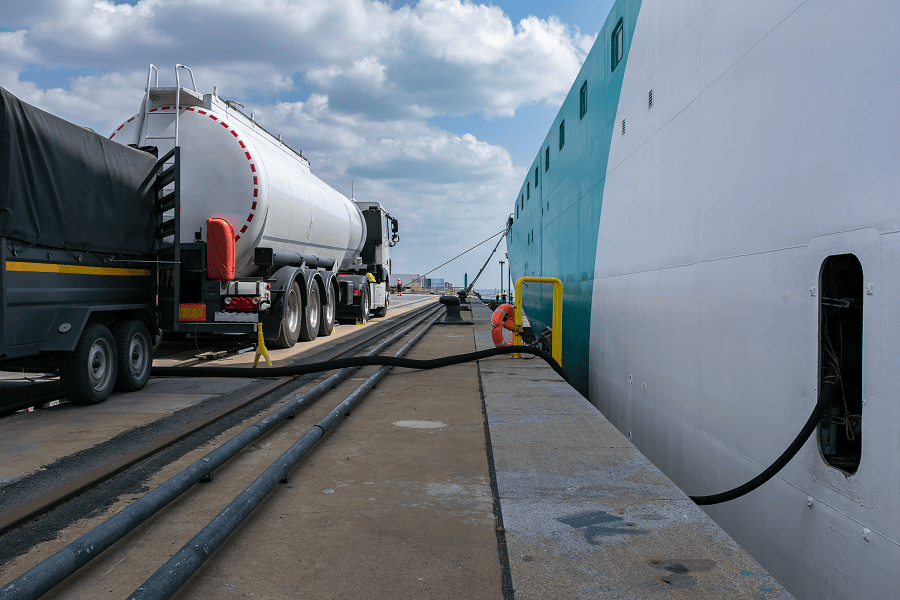 A commercial tanker truck parked at a port terminal pier actively refueling a docked maritime vessel