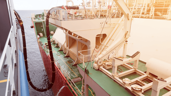 A marine ship refueling connection showing a flexible hose delivering fuel to a commercial vessel at dock