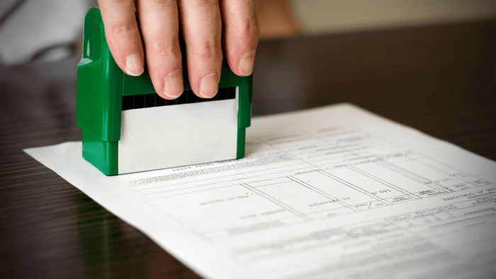 A hand uses a stamp on a document resting on a desk, representing approvals, certification, or administrative processing.