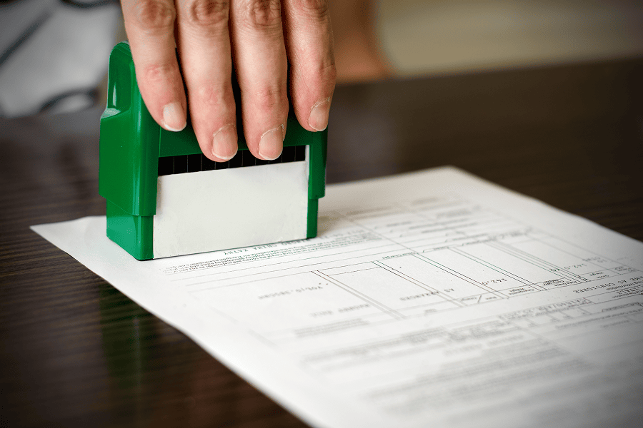 A hand uses a stamp on a document resting on a desk, representing approvals, certification, or administrative processing.