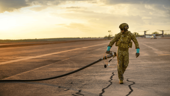 Military fuel specialist carrying a refueling hose across an airfield during aircraft fueling operations.