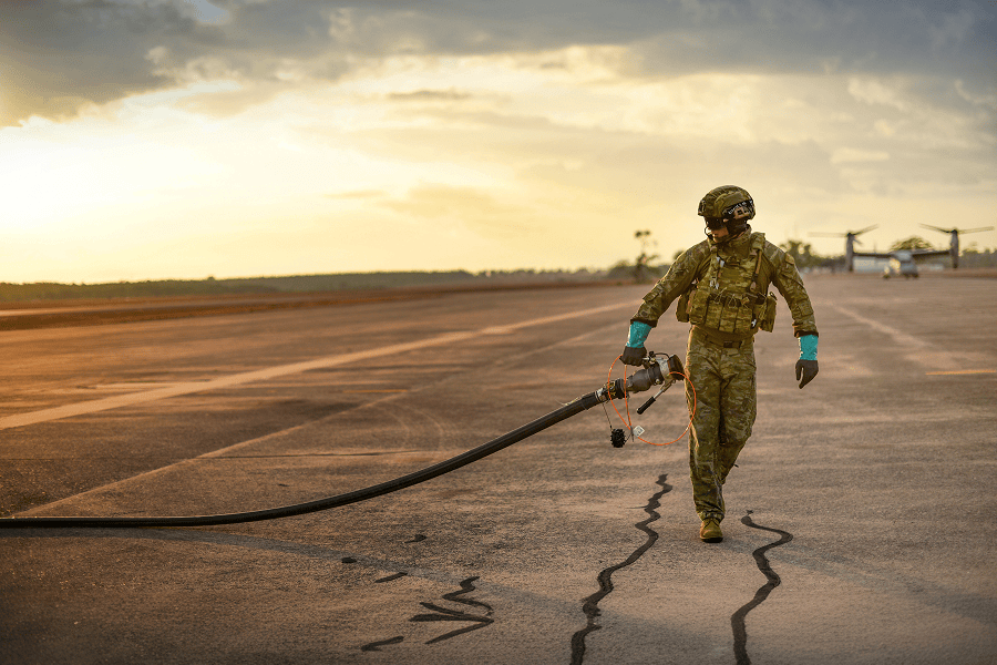 Military fuel specialist carrying a refueling hose across an airfield during aircraft fueling operations.