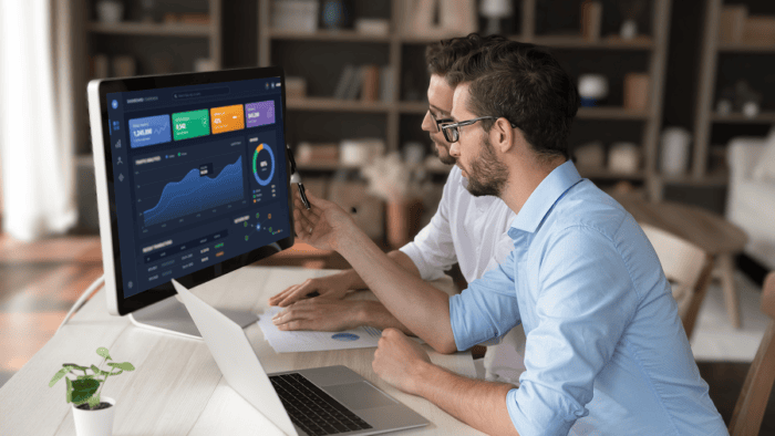 two guys at desk with market related data on screen