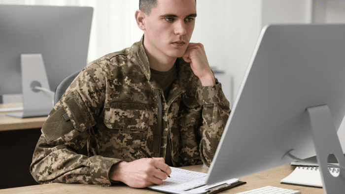 A young military soldier wearing a uniform working on a laptop at a wooden office desk