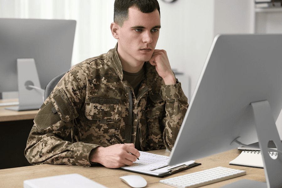 A young military soldier wearing a uniform working on a laptop at a wooden office desk